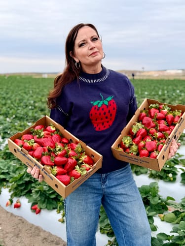 Reviewer in a strawberry field wearing the navy Strawberry Fields Sweater with jeans, holding boxes of fresh strawberries.