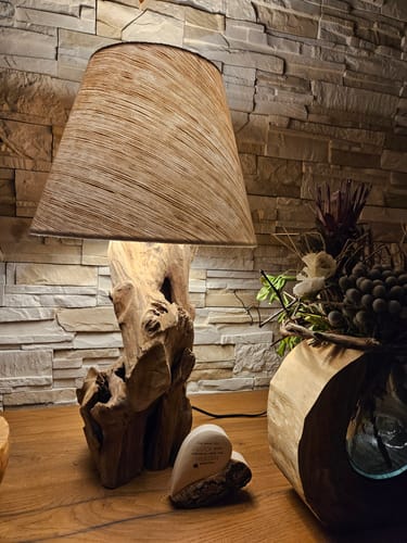 Ein Kunde zeigt die leuchtende ZOILA Treibholzlampe mit rustikalem Holzfuß auf einem Holztisch vor einer Steinwand.