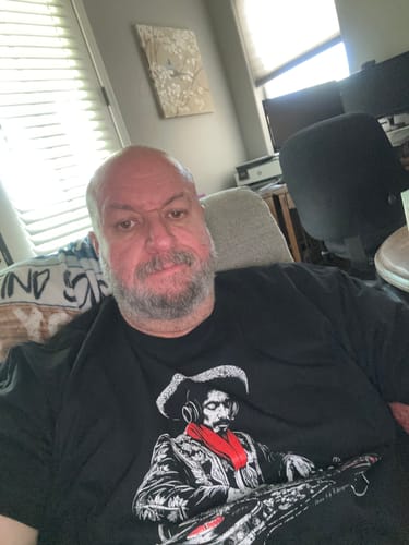Reviewer sitting indoors while wearing the black El Mariachi Ritmo Mexicano T-Shirt.