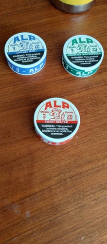 Customer's three tins of ALP nicotine pouches on a wooden table, featuring Sweet Nectar, Chilled Mint, and Mountain Wintergreen flavors.