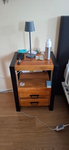 Recensent toont het Mango Houten Nachtkastje Britt in een slaapkamer, met twee lades en een zwart frame.
