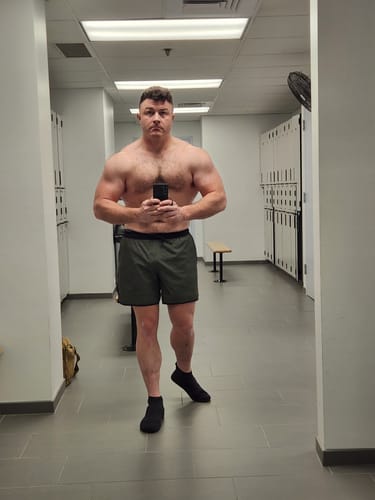 Reviewer standing in a gym locker room wearing the dark green Covert Camo – V2 Lined Training Shorts.