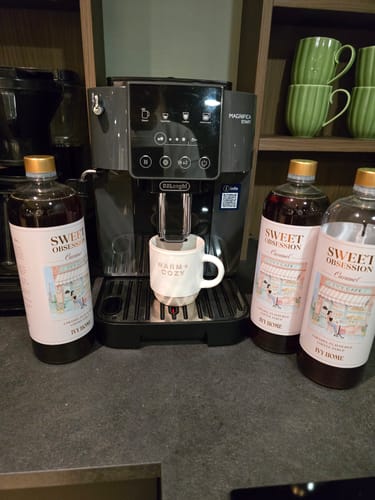 Customer's four 1L bottles of Sweet Obsession Caramel Coffee Syrup on a dark counter next to a coffee machine.