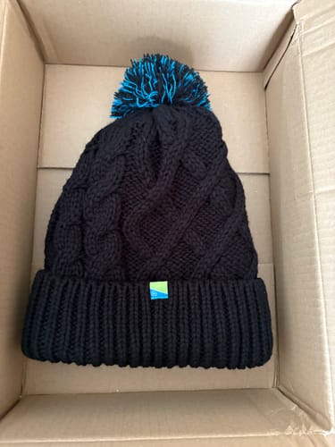 Recensent toont de zwarte gebreide pomponmuts met een blauwe pompon in een kartonnen doos.