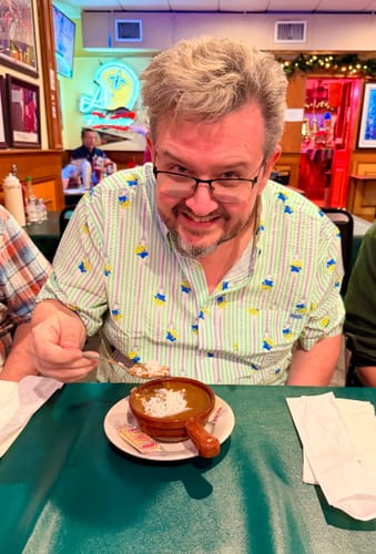 Reviewer smiling in the Toy Story 'The Claaaaaw' seersucker shirt while eating gumbo at a restaurant.