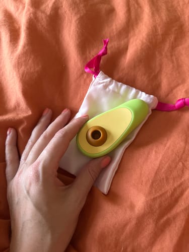 Customer's hand holding the green Avo Clitoral Massager on its white pouch against an orange background.
