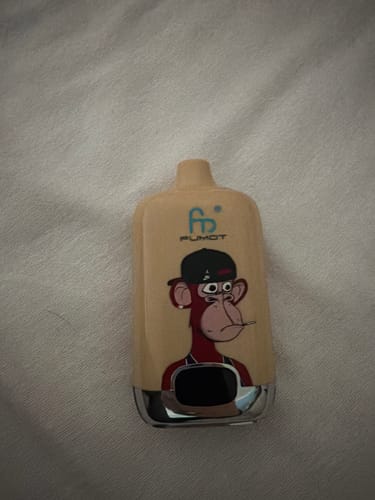 Ein Kunde zeigt die beigefarbene RANDM DIGITAL BOX 12000 Vape mit Affen-Grafik auf einer weißen Oberfläche.