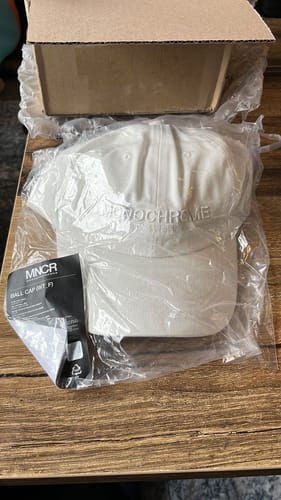 Reviewer shows a new white 'MONOCHROME' BTS baseball cap in its packaging, an item from their K-POP RANDOM BOX.
