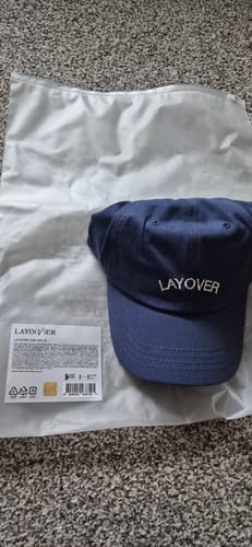 Reviewer shows a navy blue 'Layover' baseball cap received in their K-POP RANDOM BOX.