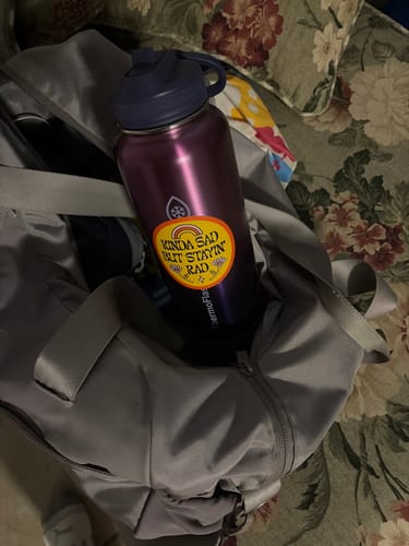 Reviewer's 'Kinda Sad But Stayin' Rad' sticker on a purple water bottle inside a grey gym bag.