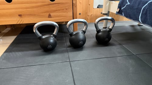 Rezensent zeigt drei aufgereihte Rubber Kettlebell PROs auf einer schwarzen Bodenmatte.