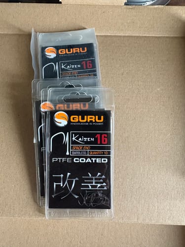 Klant toont meerdere verpakkingen van Guru Kaizen Spade End Barbless haken, maat 16, in een doos.