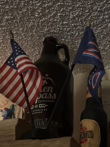 Customer's America 250™ Stick Flag Set displayed on a dark shelf next to some bottles.