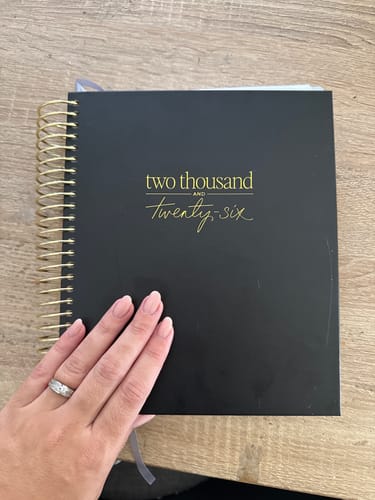 Customer holding the black 2026 Signature Planner, showing its gold spiral binding and foil text on the cover.