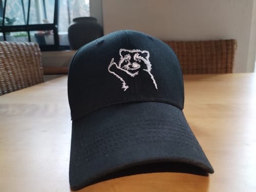 Rezensent zeigt die schwarze Baseballkappe mit dem gestickten Motiv eines frechen Waschbären.