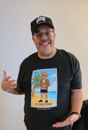 A happy reviewer wearing and gesturing towards their black El Halftime OG T-Shirt.