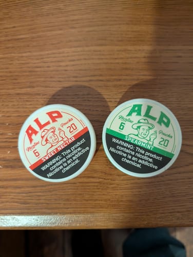Reviewer's tins of ALP Spearmint and Sweet Nectar nicotine pouches side-by-side on a wooden surface.