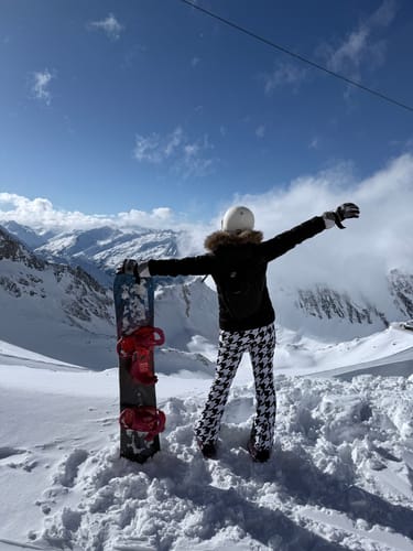 Reviewer wearing the houndstooth slim-fit ski bibs while posing on a snowy mountain with a snowboard.