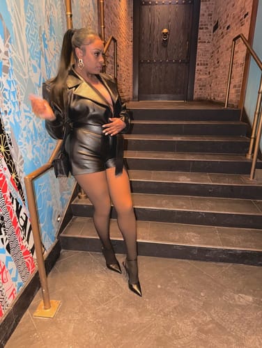 Reviewer posing on a staircase in the black Vegan Leather Trench Romper, paired with heels and pantyhose.