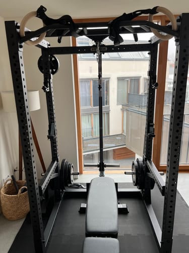 Kunde zeigt sein schwarzes R7-Cage Power Rack mit Hantelbank und Gewichten, das vor einem großen Fenster aufgestellt ist.