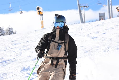 Customer skiing in the black and khaki insulated color-block one-piece ski suit on a sunny, snowy slope.