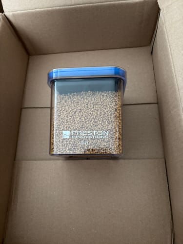 Recensent toont de Preston Bait Safe-container, gevuld met aas, in een kartonnen doos.