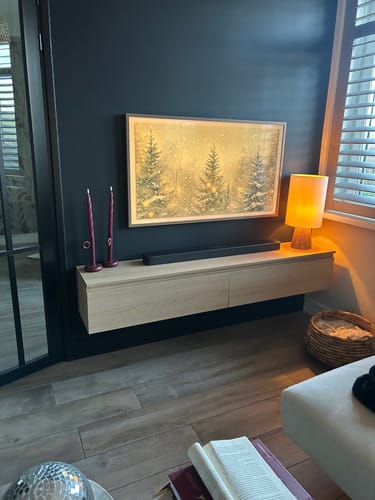 Recensent's zwevende TV-meubel Kate in een lichte kleur, gemonteerd aan een donkere muur onder een televisie.