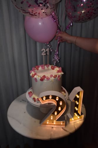 Customer's white 21st birthday cake decorated with BEGONIA edible flowers, shown with balloons and a light-up '21'.
