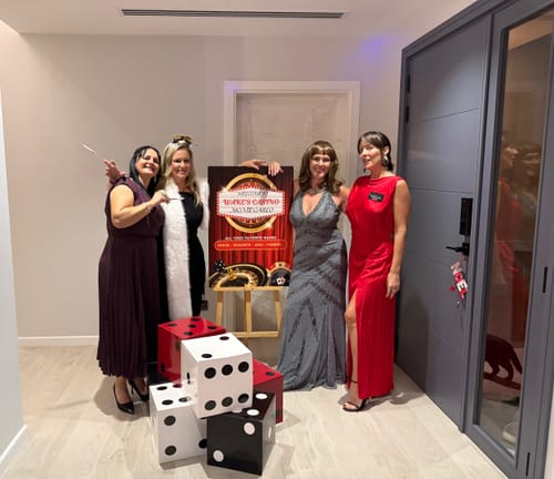 Reviewers in formal dresses posing with a poster and large dice for their Spy Murder Mystery Game party.