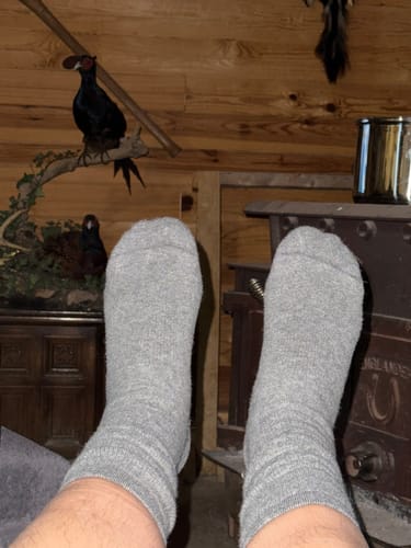 Reviewer's feet wearing grey Men's Alpaca Socks, propped up near a wood stove in a rustic room.