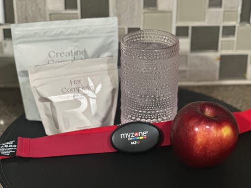 Reviewer's HER Complete supplement pouch next to an apple, a glass of water, and fitness gear.