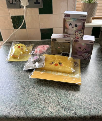 Customer's K-POP RANDOM BOX contents, including ATEEZ plush keyrings and a yellow pouch, displayed on a counter.