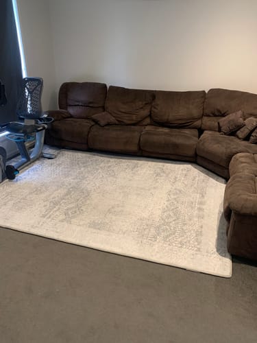 Customer's Silver Mist Mellow Mat on a dark floor in a living room, positioned in front of a large brown sectional sofa.
