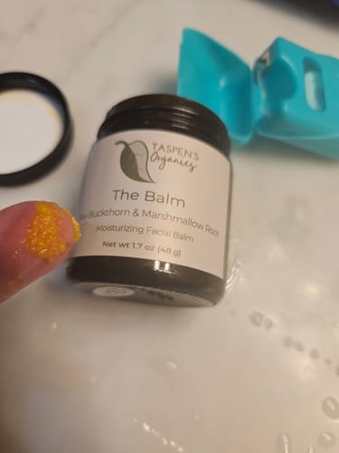 Close-up of a reviewer's fingertip with the orange-colored The Balm on it, next to the open product jar.