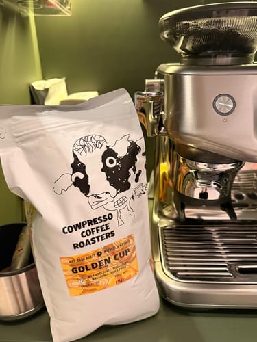 Customer's white bag of Golden Cup Blend coffee sitting on a counter beside a silver espresso machine.