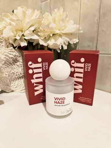 Customer's Vivid Haze perfume bottle and red boxes displayed on a white counter in front of a bouquet of white flowers.