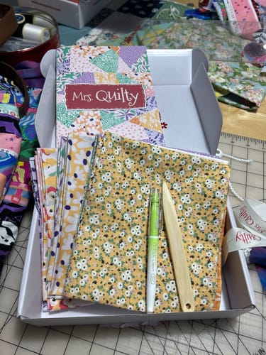 Customer's Mrs Quilty Subscription Box opened to show patterned fabrics, an instruction booklet, and sewing tools.