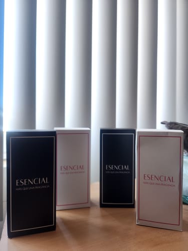 Un cliente muestra cuatro cajas de perfume Esencial, dos negras y dos blancas, colocadas sobre una mesa de madera.