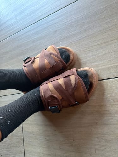 Reviewer wearing brown Chocolate Dracos slides with black socks on a wooden floor.