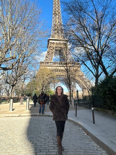 Customer wearing the chocolate faux fur jacket on a cobblestone street with the Eiffel Tower in the background.
