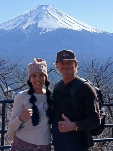 Customer wearing the Hat of the Month while posing with a companion in front of a large, snow-capped mountain.