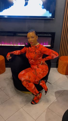 Customer sitting in a chair wearing the vibrant red Bare Body Boatneck Bodysuit and matching pants.
