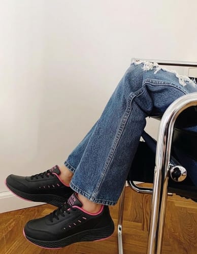 A reviewer sitting down, wearing black Soft Stride sneakers with pink trim and blue jeans.
