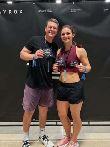 Two reviewers holding Cael's Cookies in front of a HYROX race banner.