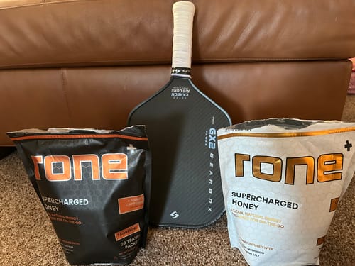 Reviewer's two bags of SuperCharged Honey, including the Tangerine flavor, next to a pickleball paddle.
