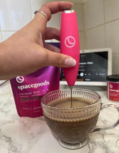 Customer mixes a frothy cup of Rainbow Dust in a mug using a pink whisk, with the product pouch behind.
