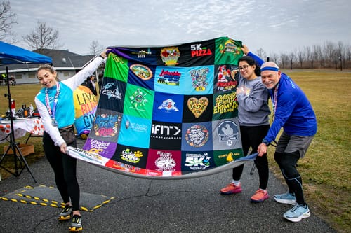 Reviewer and two others hold up a large, colorful Premium T Shirt Quilt made from various race shirts outdoors.