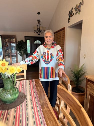 Reviewer smiling in her dining room while wearing the colorful Flores Y Corazon Art De La Calle Hoodie.