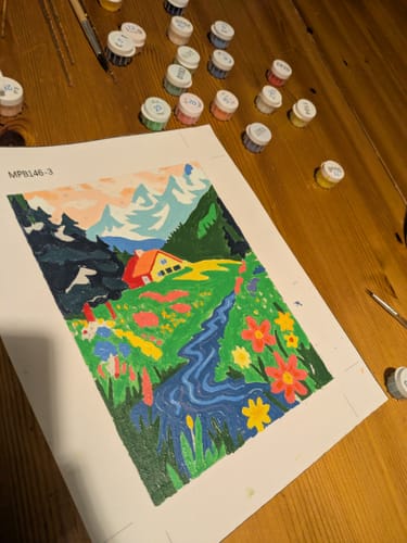 Customer's nearly finished colorful landscape from the Mini Paintings set, with paint pots and brushes on a table.