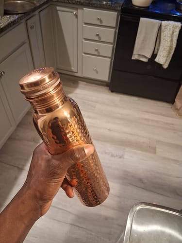 A reviewer's hand holding the shiny, hand-hammered Himalayan Ayurvedic Pure Copper Bottle in a kitchen.